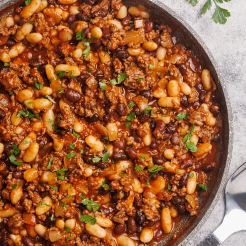 A large pan filled with baked beans and beef next to large spoon.
