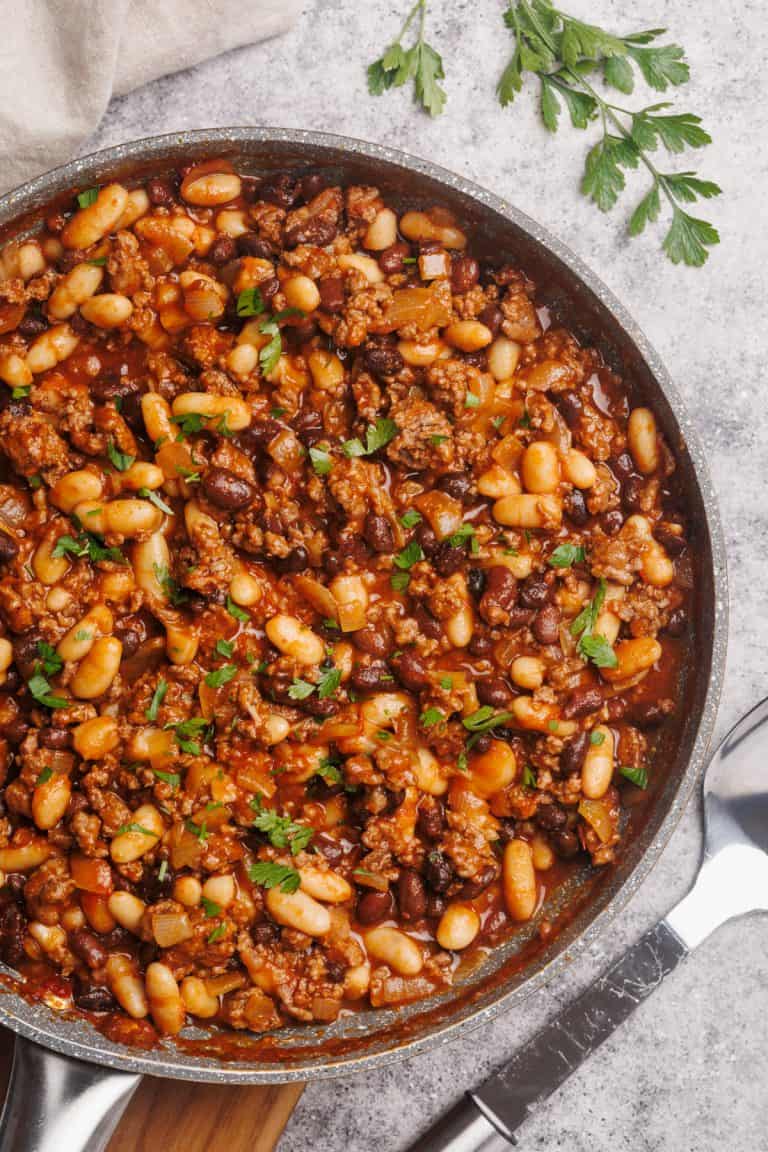 A large pan filled with baked beans and beef next to large spoon.
