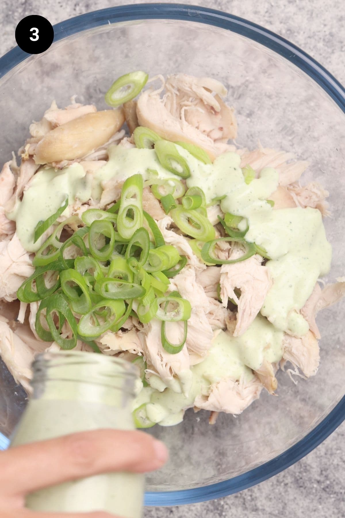 Ranch dressing being poured into a glass bowl with cooked chicken and green onions.