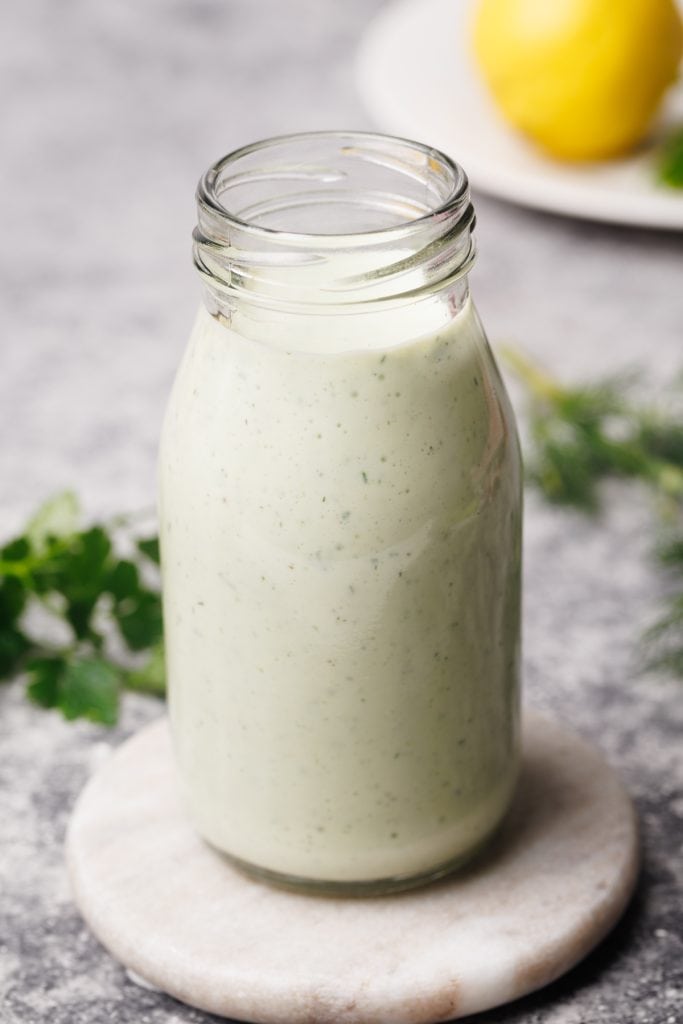 A small glass bottle filled with Greek yogurt ranch dressing on a marble coaster next to fresh herbs and a linen tea towel.
