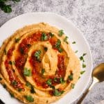 Harissa hummus on a white plate next to fresh herbs and a linen napkin.