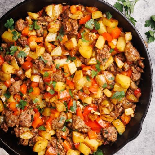 Sausage and potato hash in a cast iron pan next to a linen towel and fresh herbs.